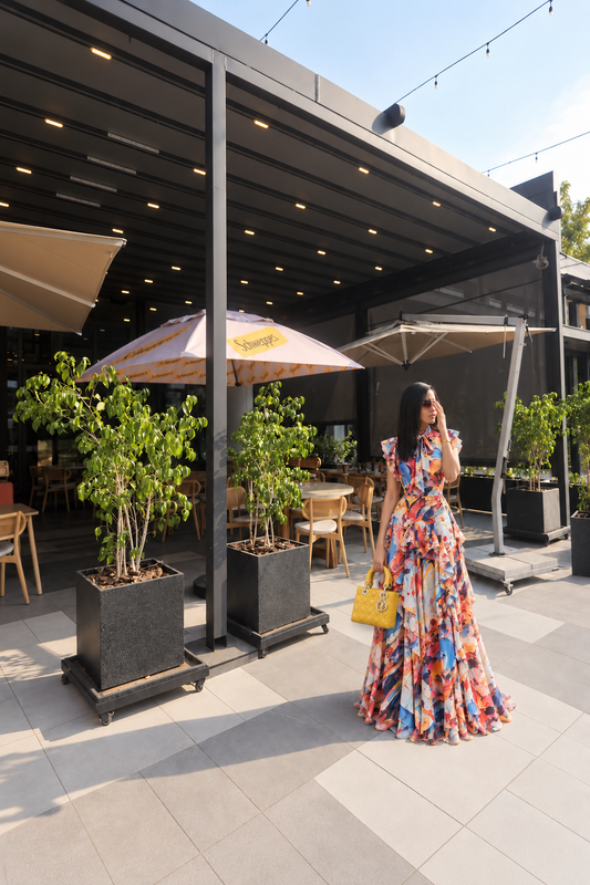 A woman wearing a vibrant, multicolored floral print maxi dress stands on an outdoor patio next to a modern cafe. She is holding a small yellow handbag.