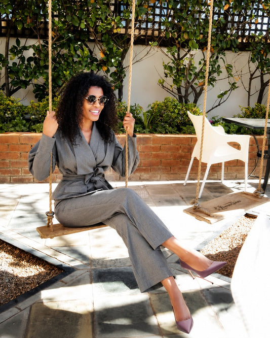 Woman in a gray outfit sitting on a swing in an outdoor setting with greenery and a white chair.
