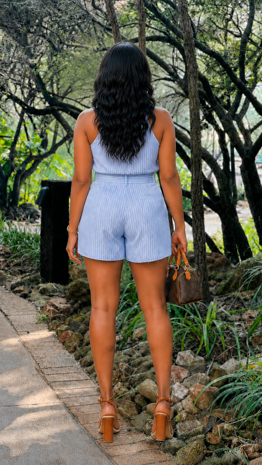 A woman viewed from behind wearing a light blue and white vertically striped sleeveless romper, walking on a stone path in a wooded outdoor setting. She is holding a brown handbag and wearing brown high-heeled sandals.