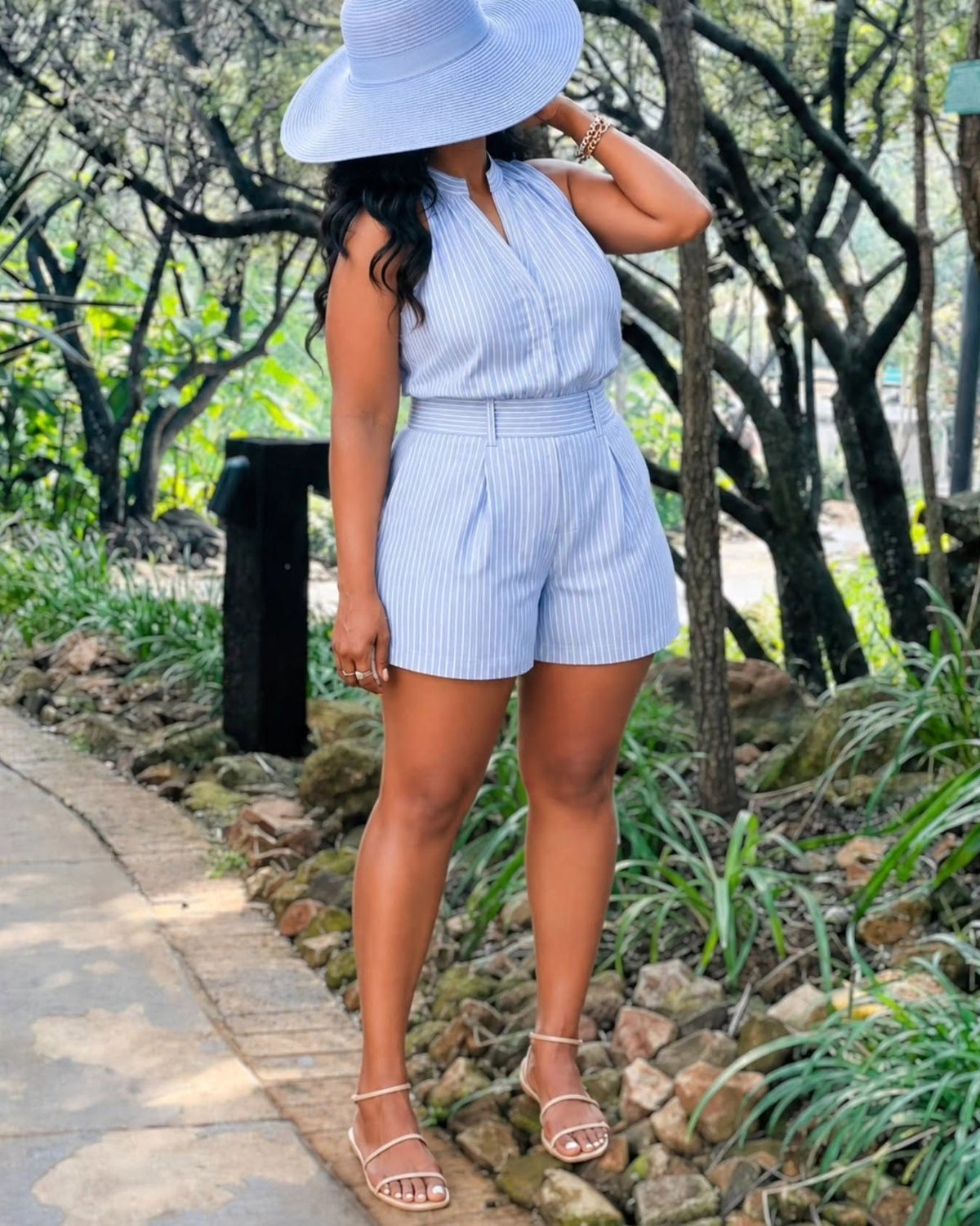 A woman wearing a light blue and white vertically striped sleeveless romper with matching shorts and a wide-brimmed light blue hat, standing outdoors on a stone path surrounded by trees and greenery.