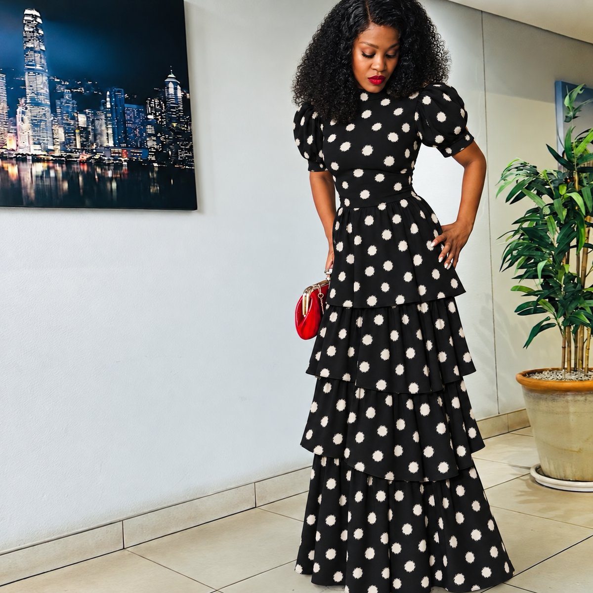 A woman wearing a black midi dress with white polka dots, puff sleeves, and a tiered ruffled hem, holding a small red handbag in an indoor setting.