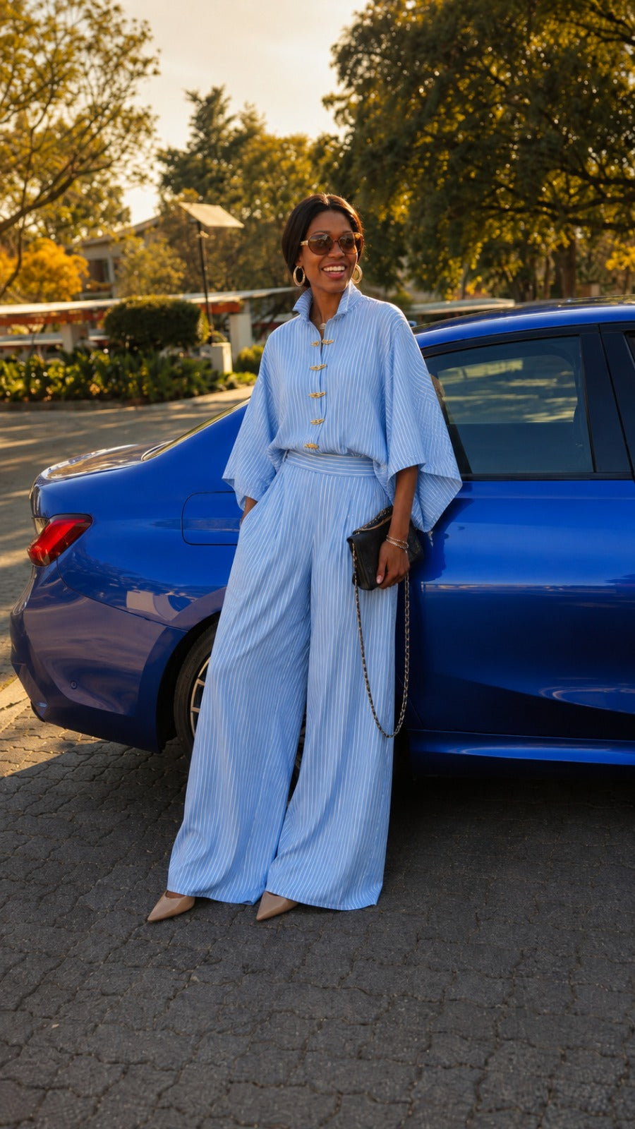 A woman wearing a light blue striped button-up jumpsuit with wide sleeves and matching wide-leg pants stands next to a blue car on a paved street at sunset.