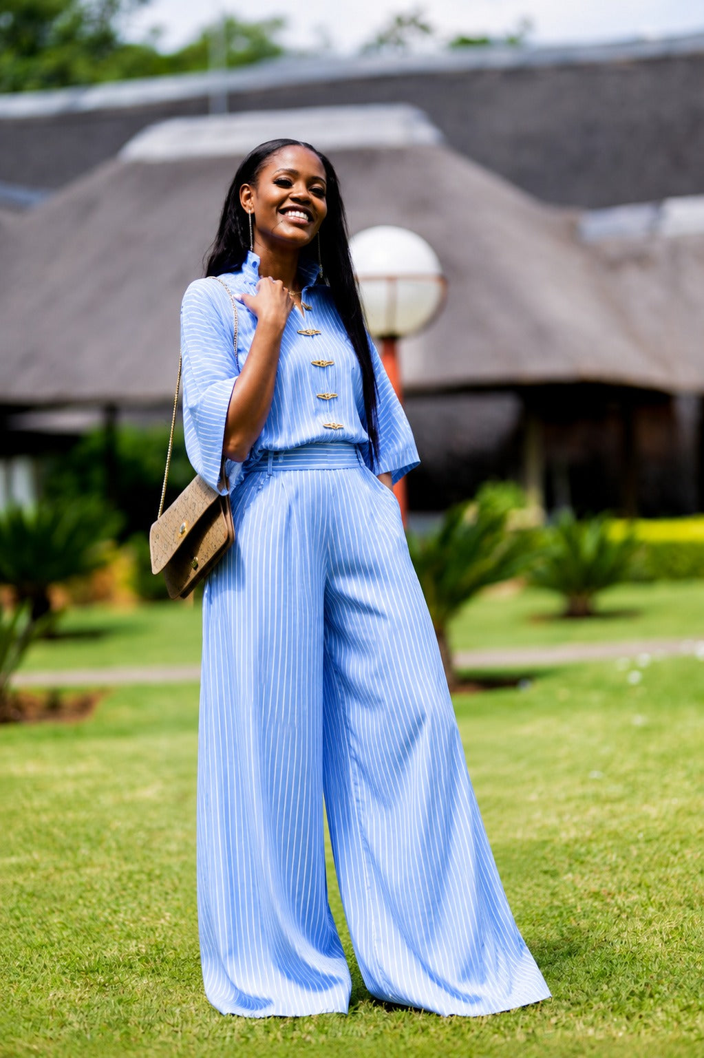 A woman stands on a grassy lawn wearing a light blue pinstriped jumpsuit with gold buttons. She is holding a small brown shoulder bag, with a modern building and trees in the background.