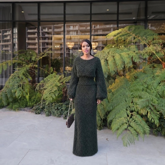 Elegant woman in dark green knit dress with long sleeves and fitted waist, standing outdoors in front of lush green plants and glass building