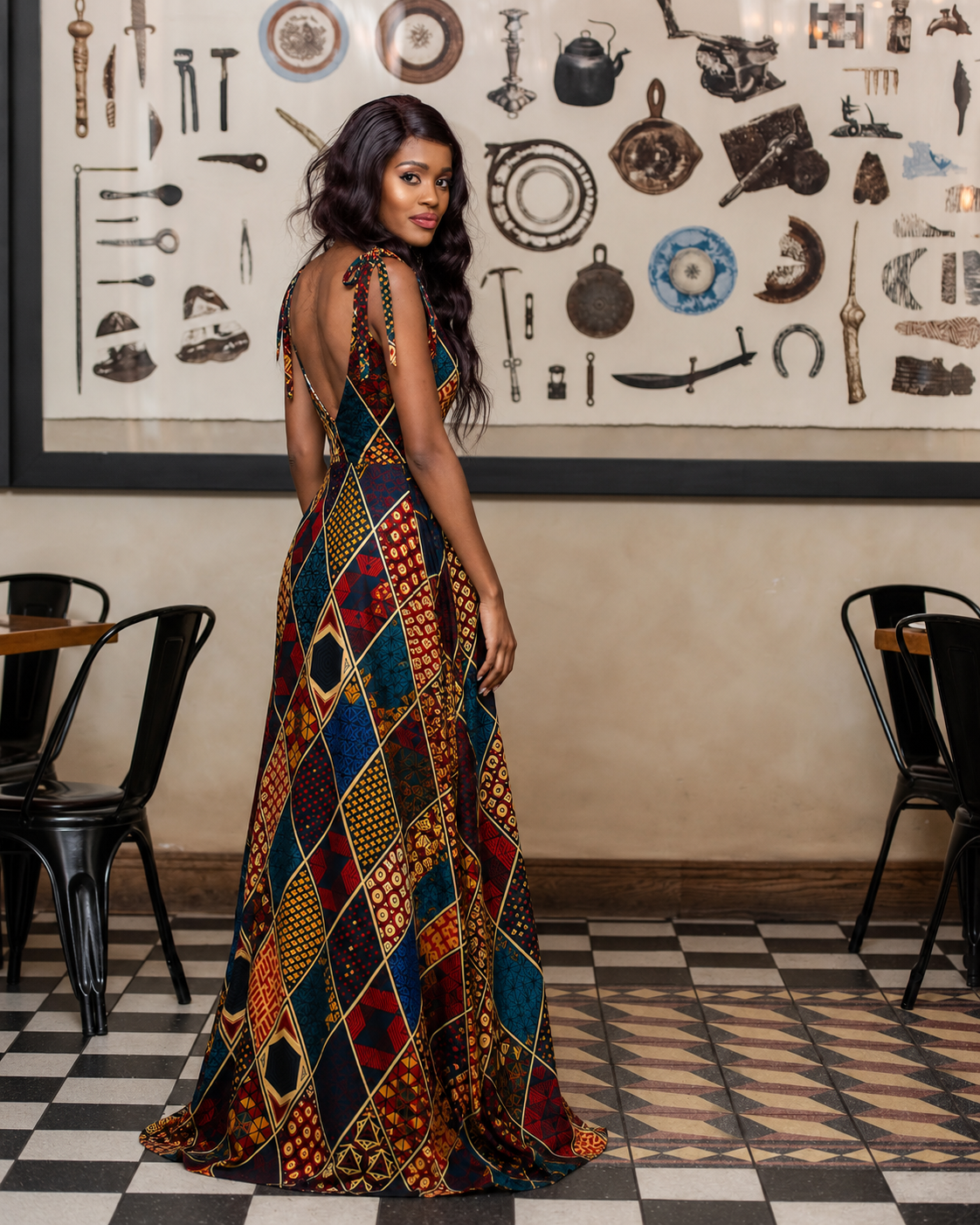 A woman stands in a restaurant wearing a sleeveless, backless A-line maxi dress with a vibrant multicolor African wax print featuring diamond patterns in red, blue, yellow, and black.