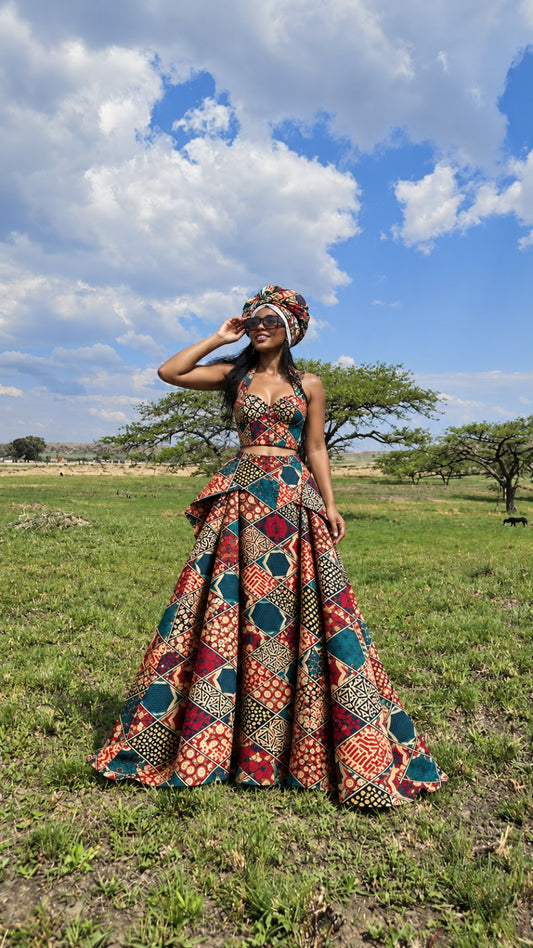 A woman wearing a vibrant, multicolored African wax print two-piece set with a matching headwrap stands in a grassy field under a blue sky with clouds.