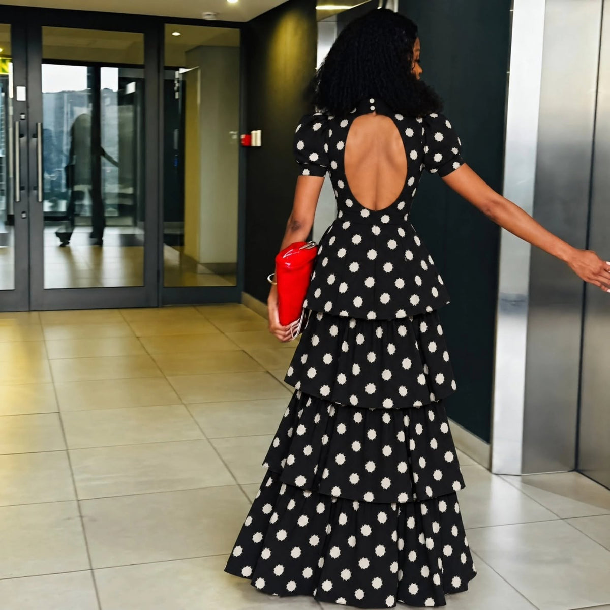 A woman wearing a black and white polka dot tiered maxi dress with a short back, walking away in a modern indoor lobby while holding a red clutch.